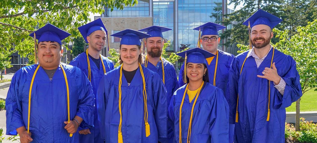 A group of graduates in blue caps and gowns pose in the campus commons